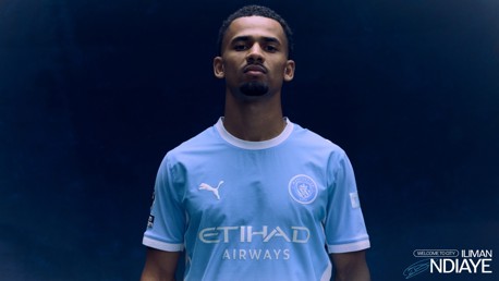 A person wearing a Manchester City football kit stands against a dark background. The kit includes the Manchester City logo and sponsors' logos.