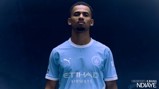 A person wearing a Manchester City football kit stands against a dark background. The kit includes the Manchester City logo and sponsors' logos.