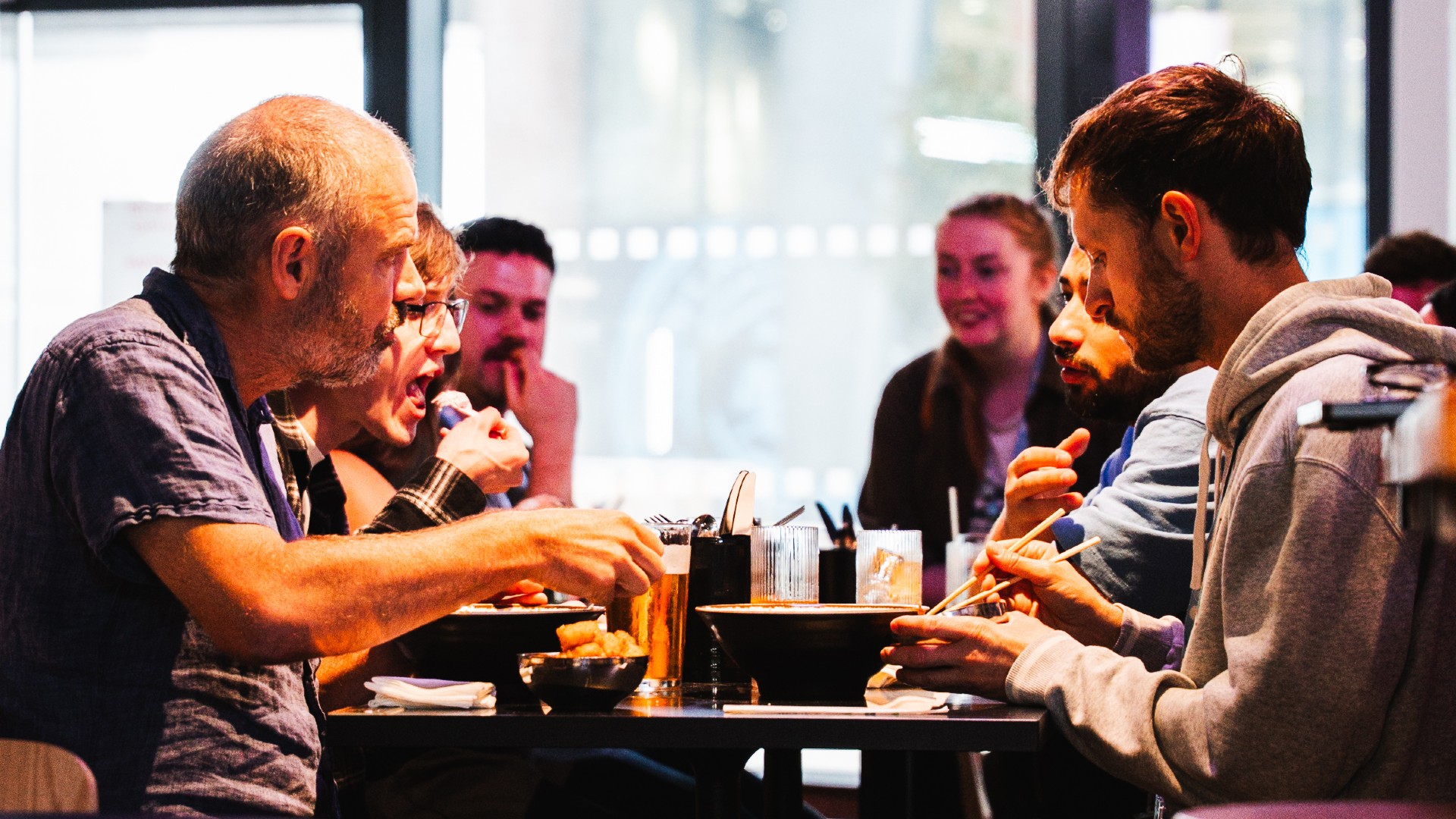 A group of people are seated at a restaurant table enjoying a meal together. They are engaging in conversation and eating with chopsticks, with drinks on the table.