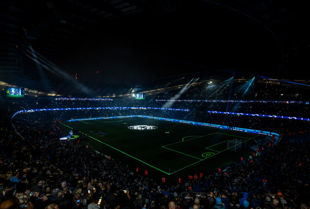 Nighttime view of a packed Etihad Stadium during a UEFA Champions League match with the field illuminated and spectators in the stands.