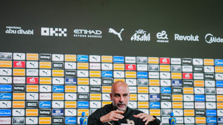 A press conference setting with a blurred person at a desk and a backdrop featuring various sponsor logos such as Etihad, Puma, Asahi, and Revolut.