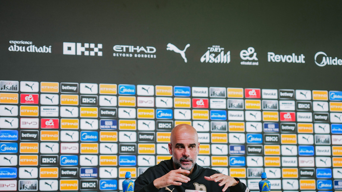 A press conference setting with a blurred person at a desk and a backdrop featuring various sponsor logos such as Etihad, Puma, Asahi, and Revolut.