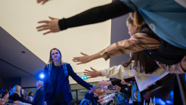 Person walking through a hallway high-fiving a line of fans, with bright lighting in the background.