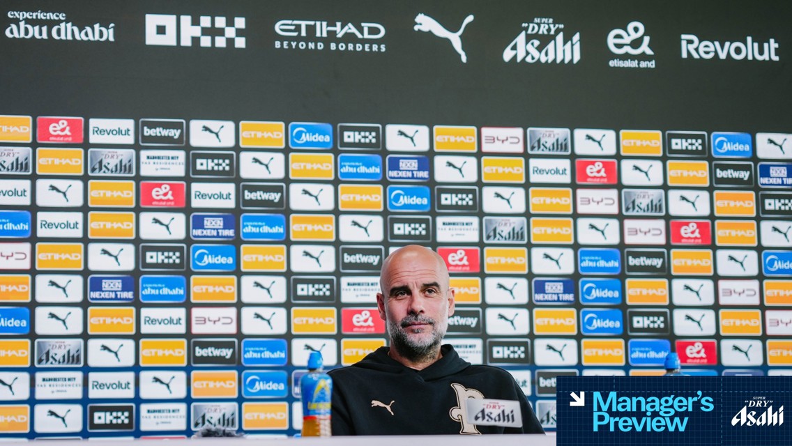A football press conference with a blurred person in front of a backdrop featuring various sponsors like Puma, Etihad, and Revolut.