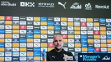 A football press conference with a blurred person in front of a backdrop featuring various sponsors like Puma, Etihad, and Revolut.