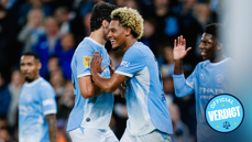 Manchester City players celebrate during a match. The players wear light blue kits, and one of them has a specific patch on the sleeve. The scene seems joyful and includes some high-fiving.