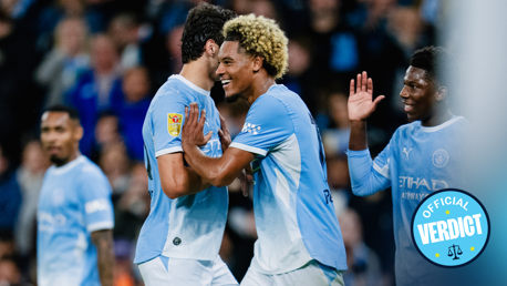 Manchester City players celebrate during a match. The players wear light blue kits, and one of them has a specific patch on the sleeve. The scene seems joyful and includes some high-fiving.