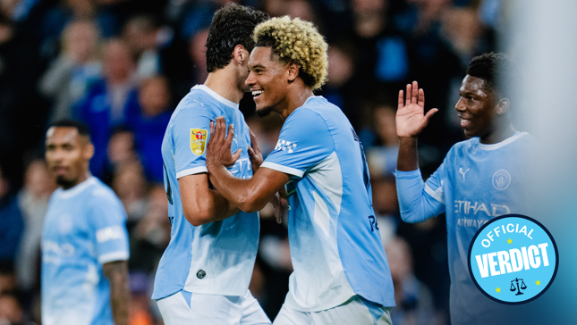 Manchester City players celebrate during a match. The players wear light blue kits, and one of them has a specific patch on the sleeve. The scene seems joyful and includes some high-fiving.