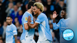 Manchester City players celebrate during a match. The players wear light blue kits, and one of them has a specific patch on the sleeve. The scene seems joyful and includes some high-fiving.