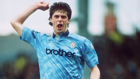 Blurred image of a football player in a classic Manchester City kit celebrating with his fist raised. The jersey features 'brother' branding and the iconic club crest on the chest.