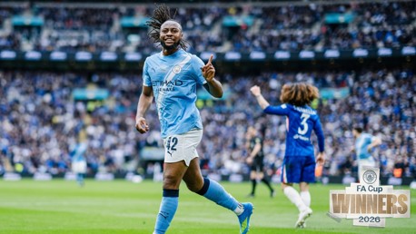 A football player from Manchester City celebrating on the field with a Chelsea player in the background. The scene is from the 2026 FA Cup final.