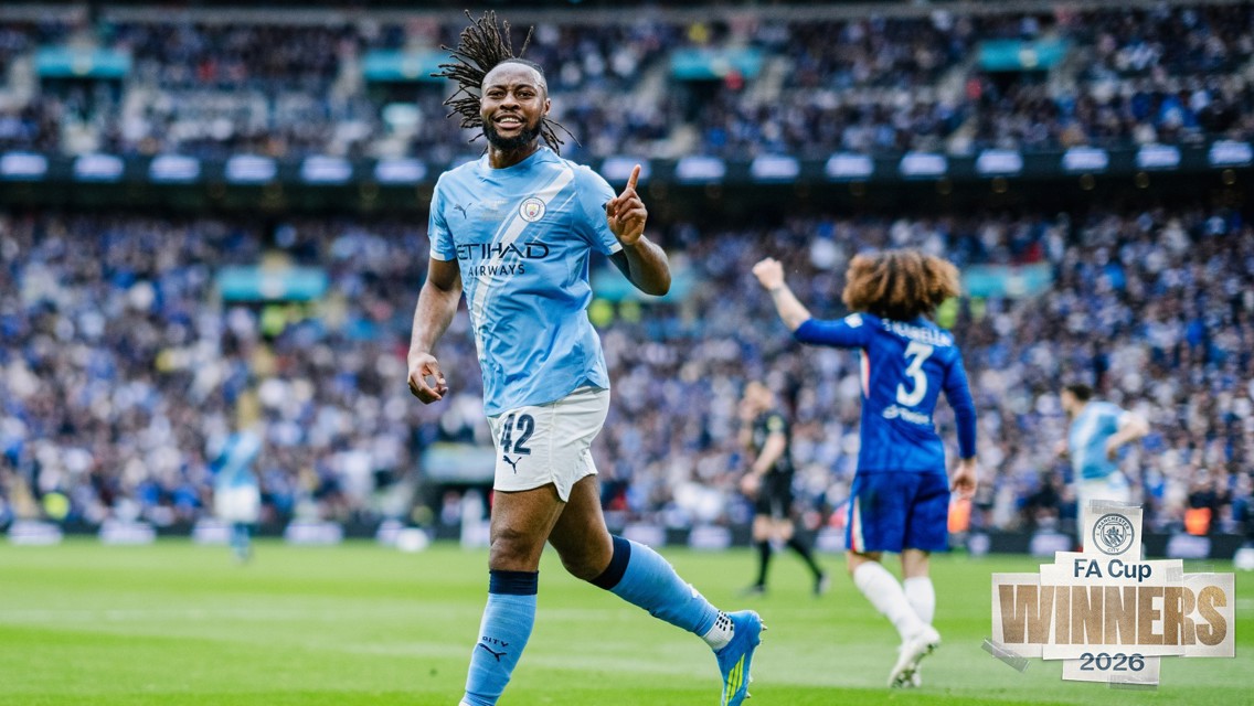 A football player from Manchester City celebrating on the field with a Chelsea player in the background. The scene is from the 2026 FA Cup final.
