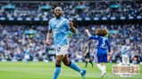 A football player from Manchester City celebrating on the field with a Chelsea player in the background. The scene is from the 2026 FA Cup final.