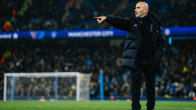 A football coach is standing on the pitch, pointing towards the field. The setting includes Manchester City branding and a stadium crowd in the background.