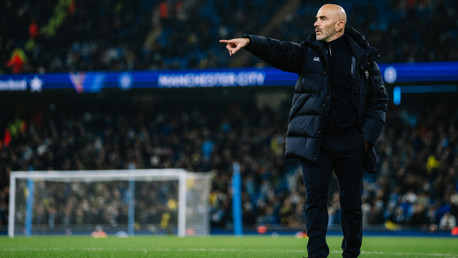 A football coach is standing on the pitch, pointing towards the field. The setting includes Manchester City branding and a stadium crowd in the background.