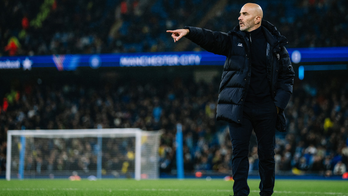 A football coach is standing on the pitch, pointing towards the field. The setting includes Manchester City branding and a stadium crowd in the background.