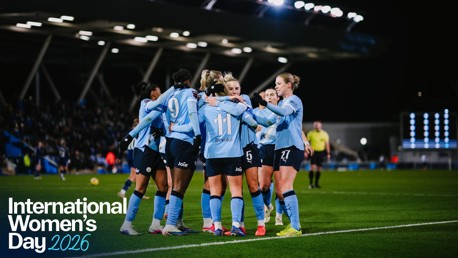 Manchester City women's soccer team celebrates as a group on the field during a match at night. The image promotes International Women's Day 2026.