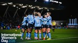 Manchester City women's soccer team celebrates as a group on the field during a match at night. The image promotes International Women's Day 2026.
