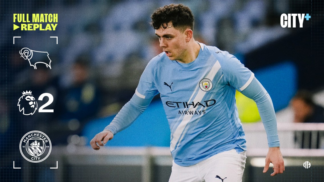 A Manchester City player in a light blue jersey during a match against Derby County. The score shown is Manchester City 2, Derby County 0.