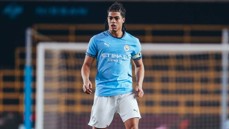 A football player wearing a Manchester City jersey is standing on the pitch with a blurred background of a goal's net.