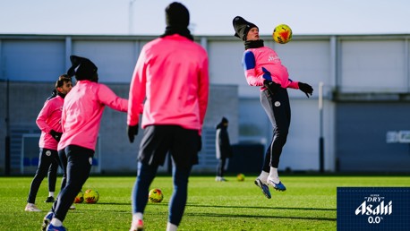 A group of football players in pink training gear practice on a field. One player is jumping to head a ball while others observe.