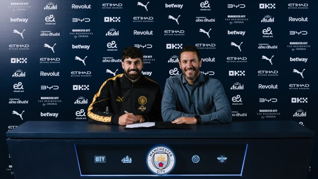 Two individuals sitting at a table with Manchester City branding. One is signing a document. Logos from various sponsors are visible in the background.