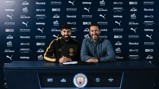 Two individuals sitting at a table with Manchester City branding. One is signing a document. Logos from various sponsors are visible in the background.