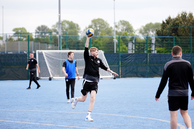 A group of players engaged in a Manchester City training session on a blue field; one player is leaping to catch a ball.