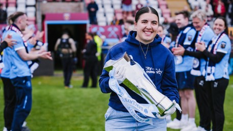 A person in a blue hoodie holding a silver football trophy on a field, surrounded by people clapping and wearing light blue shirts and scarves.