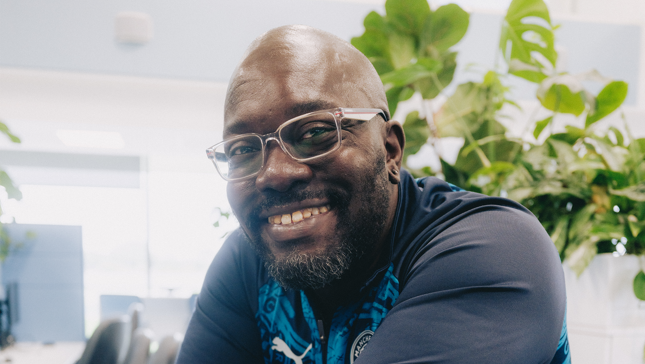 The person is smiling at a desk, wearing glasses and a blue Manchester City top, surrounded by plants.