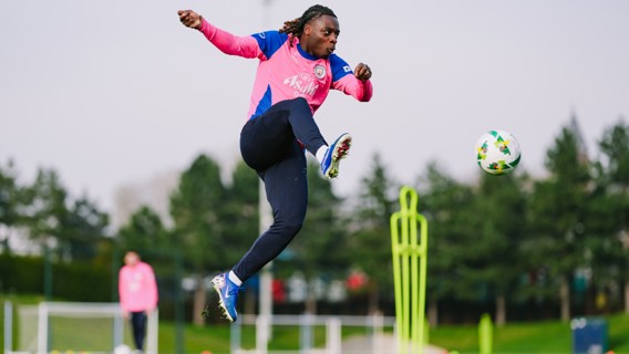 A person in a pink and blue sports outfit performs a high kick towards a soccer ball during practice on a field with cones and training aids.