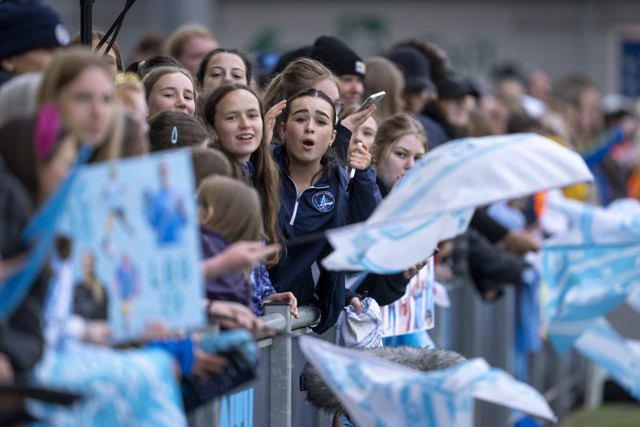 A group of enthusiastic fans, mostly young people, are gathered behind a barrier at a sporting event, waving blue and white flags and holding signs.