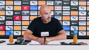 A person at a press conference with a Manchester City backdrop, microphones, and Lucozade bottles on the table.