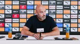 A person at a press conference with a Manchester City backdrop, microphones, and Lucozade bottles on the table.