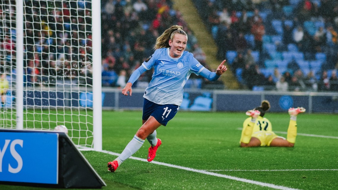 A female Manchester City football player in a light blue jersey celebrating a goal, running near the goalpost. Another player in a yellow kit is on the ground in the background.