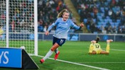 A female Manchester City football player in a light blue jersey celebrating a goal, running near the goalpost. Another player in a yellow kit is on the ground in the background.