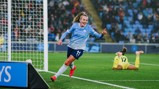 A female Manchester City football player in a light blue jersey celebrating a goal, running near the goalpost. Another player in a yellow kit is on the ground in the background.