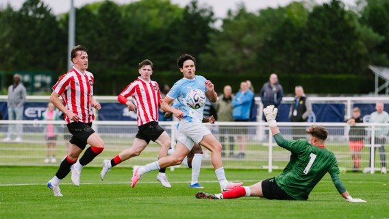 Soccer players in action during a youth match. A player in a light blue kit is attempting to score past a goalkeeper in green, with defenders in red and white striped kits running towards them on a green field.