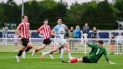 Soccer players in action during a youth match. A player in a light blue kit is attempting to score past a goalkeeper in green, with defenders in red and white striped kits running towards them on a green field.
