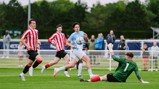 Soccer players in action during a youth match. A player in a light blue kit is attempting to score past a goalkeeper in green, with defenders in red and white striped kits running towards them on a green field.