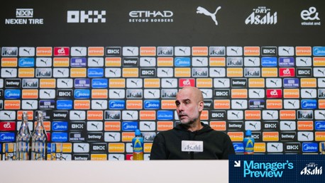 A press conference backdrop featuring Manchester City logos and sponsors like Nexen Tire, OKX, Puma, Etihad. Bottles and a 'Manager's Preview' sign are on the table.