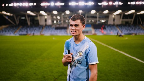 A person in a Manchester City jersey holding a microphone on a football field in a stadium setting.