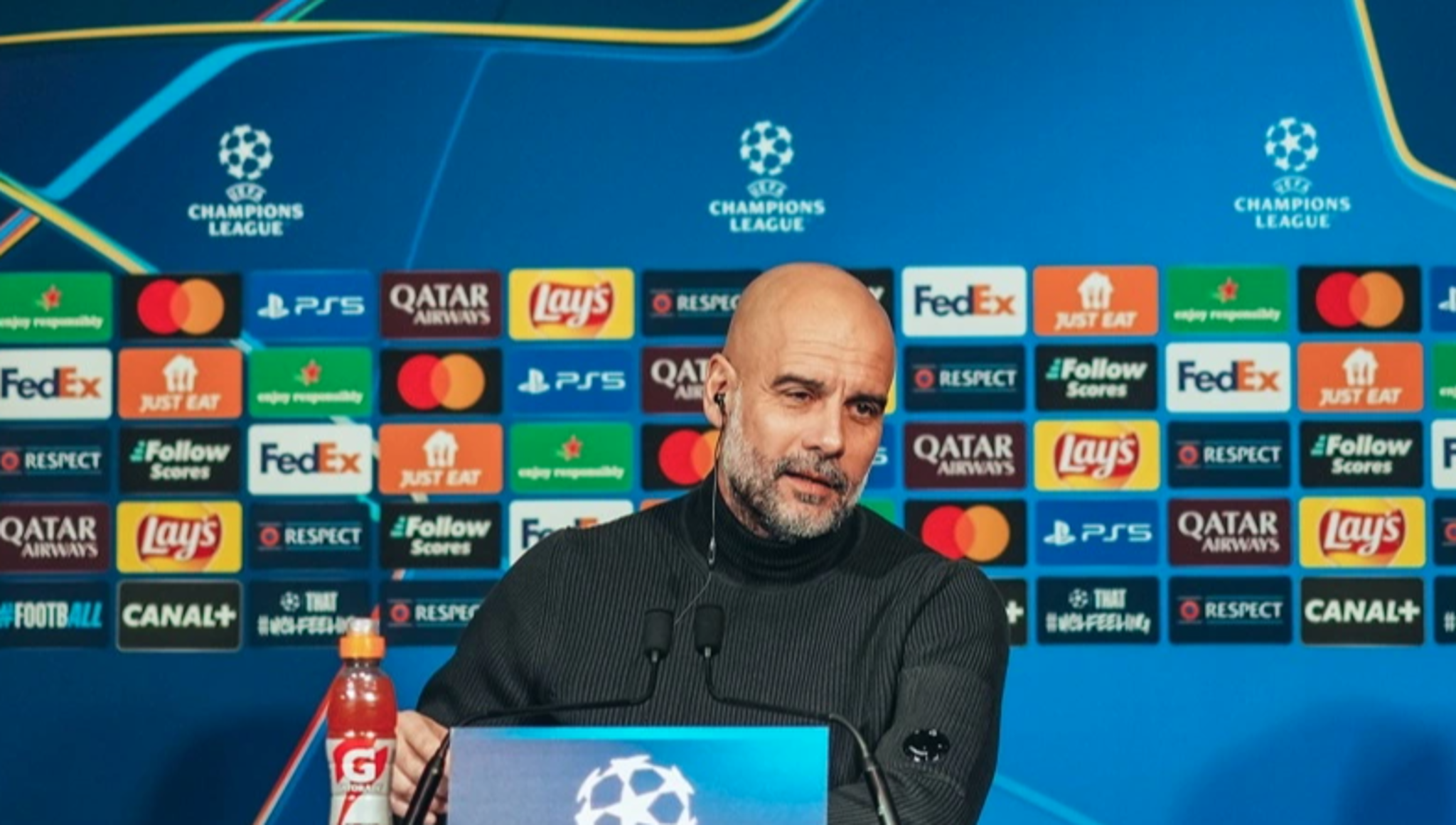A press conference scene with a person sitting behind a desk and in front of a backdrop featuring various sponsor logos, including Lay's, FedEx, and Qatar Airways, associated with the UEFA Champions League.