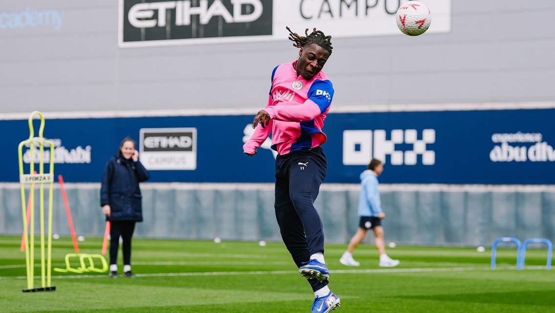 A soccer player in pink and blue training attire is jumping to head a ball during practice at the Etihad Campus, with others and equipment visible in the background.
