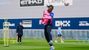 A soccer player in pink and blue training attire is jumping to head a ball during practice at the Etihad Campus, with others and equipment visible in the background.