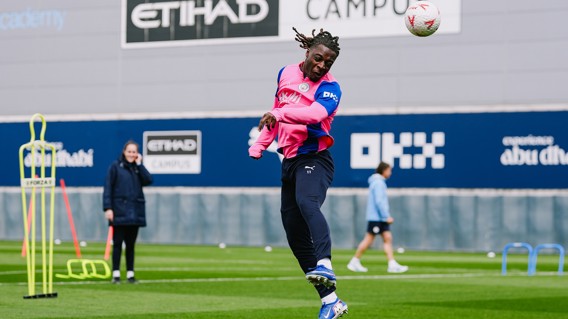 A soccer player in pink and blue training attire is jumping to head a ball during practice at the Etihad Campus, with others and equipment visible in the background.