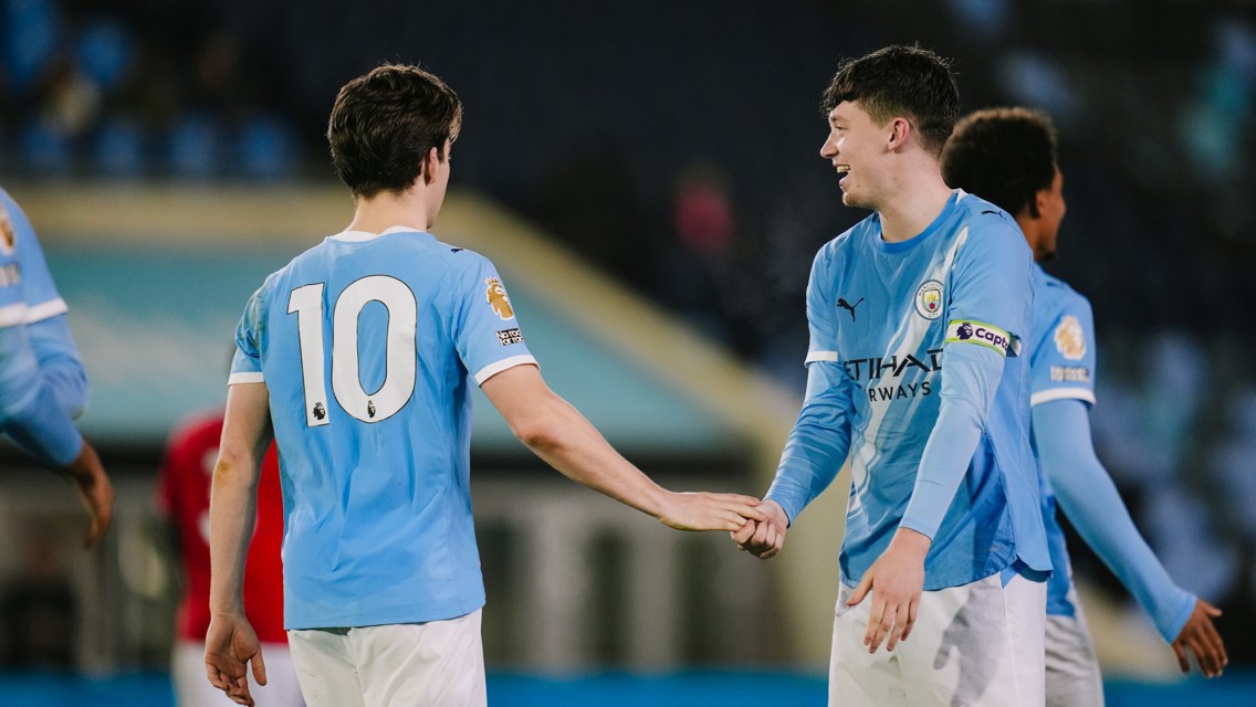 Two Manchester City players in blue jerseys shaking hands on the pitch. One player wears a captain's armband.