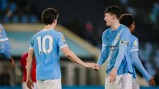 Two Manchester City players in blue jerseys shaking hands on the pitch. One player wears a captain's armband.