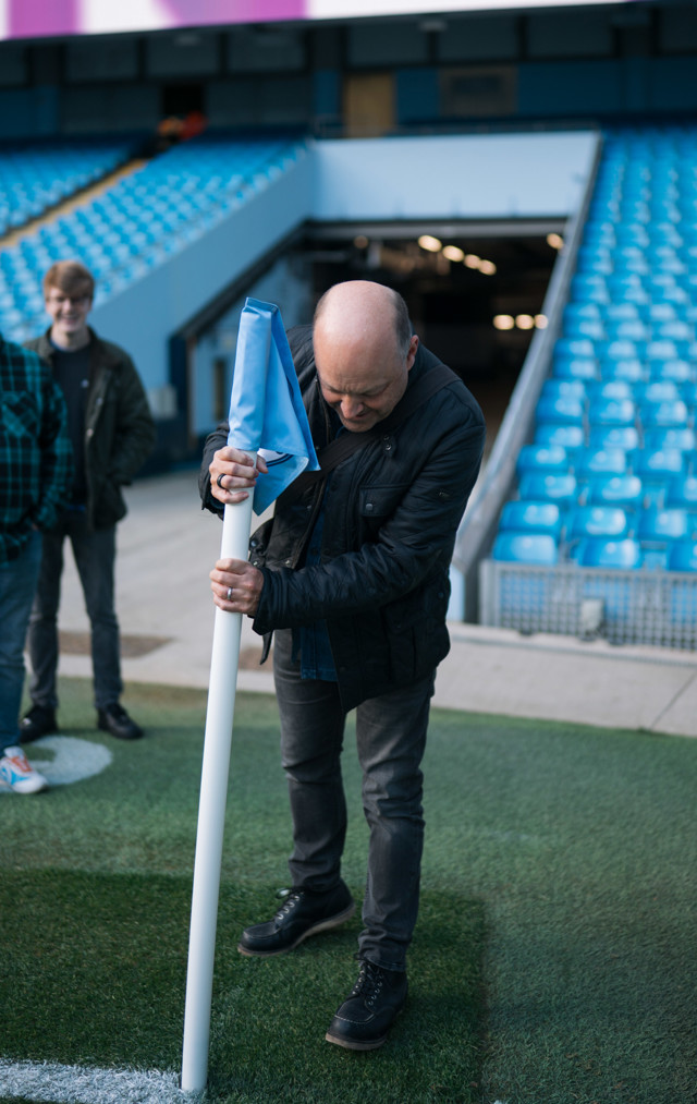A man in a black jacket adjusts a blue corner flag on a football field. He is standing near the sideline in a stadium with blue seats.