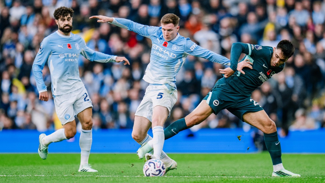 Players from Manchester City and Brighton compete for the ball during a match. Two Manchester City players in light blue jerseys challenge a Brighton player in dark jersey.